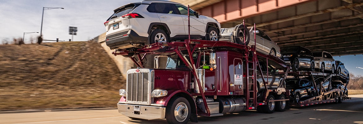 Burgundy truck transporting cars under overpass.