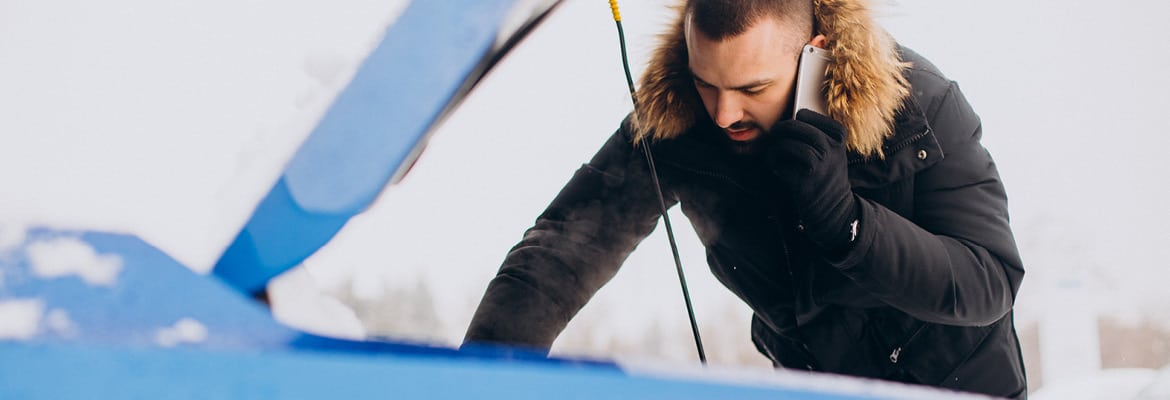 A man in winter clothes holding a cell phone to his ear while looking in the hood of a car