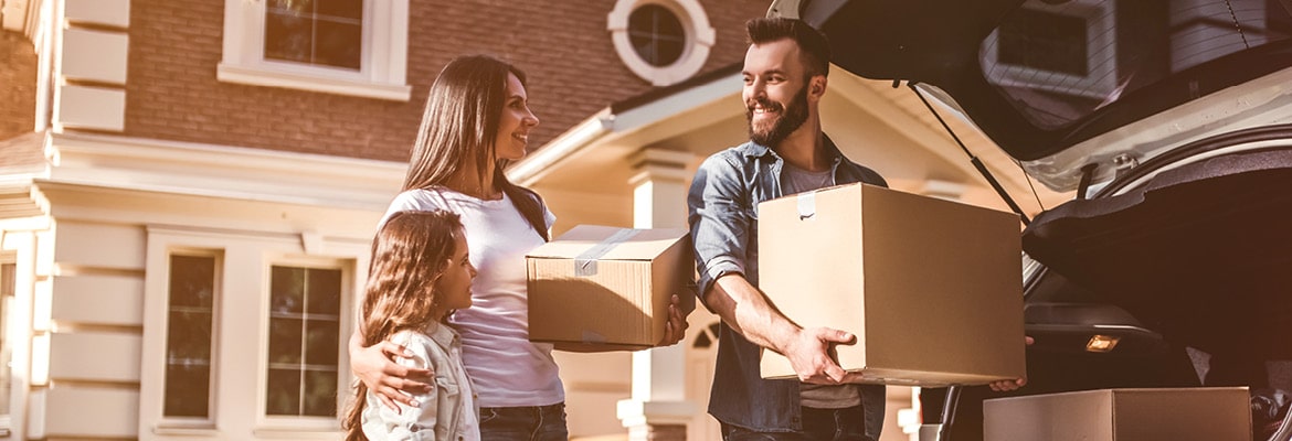 A family standing outside of a house carrying moving boxes out of their car.