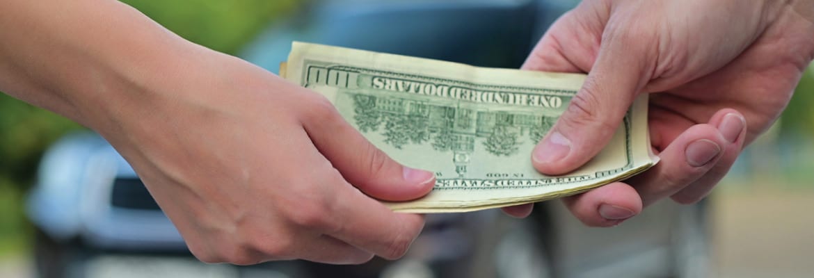 Two hands exchanging cash in front of a car