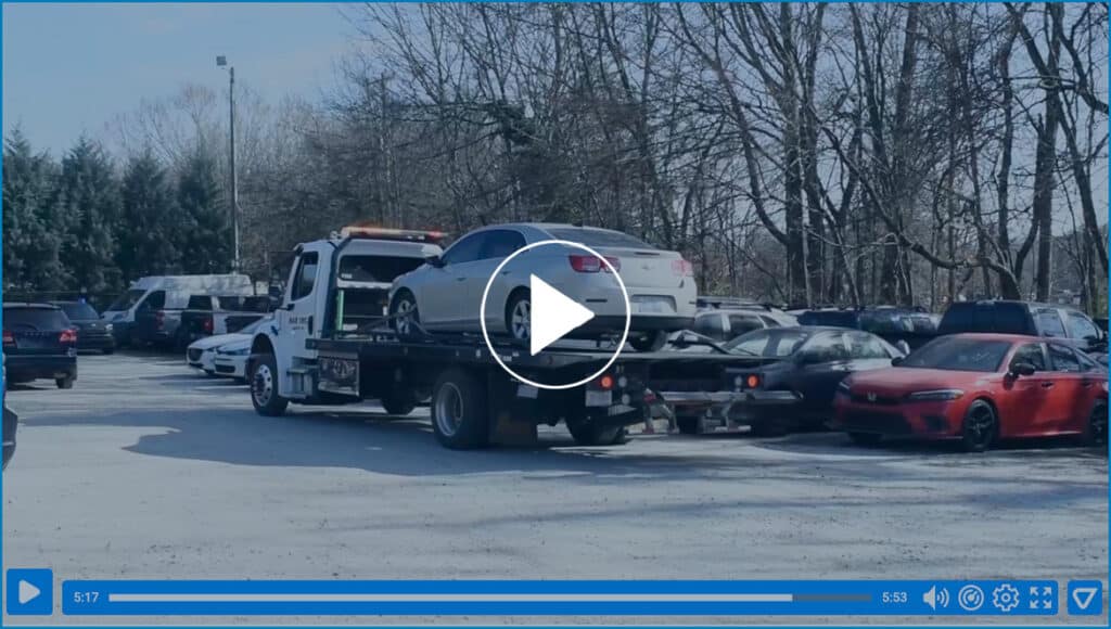 Video screenshot with a play button. Tow truck hauling a car in the background.