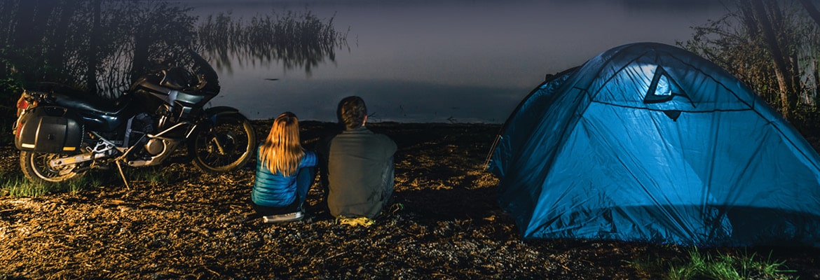 Man and woman sitting between blue tent and motorcycle while camping.