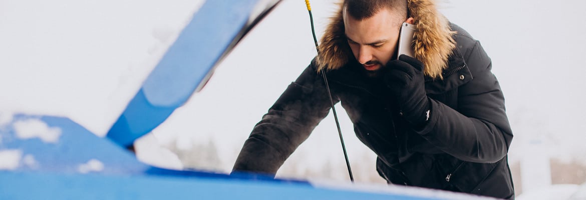 Man in winter coat talks on phone while looking under hood of car.
