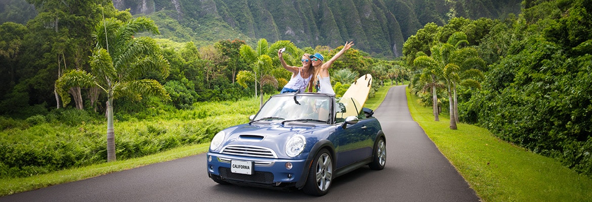 Two women driving convertible car in Hawaii with a surfboard.